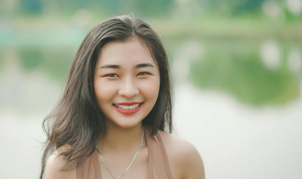 Smiling young woman with long hair, showcasing a bright smile by a serene water backdrop, reflecting confidence and beauty, relevant to cosmetic dentistry in Anaheim.