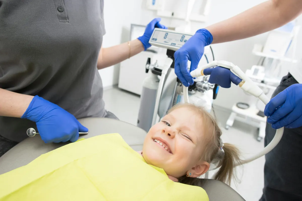 Child smiling in a dental chair, wearing a yellow bib, with dental professionals in blue gloves preparing for a pediatric dental procedure in a child-friendly environment.